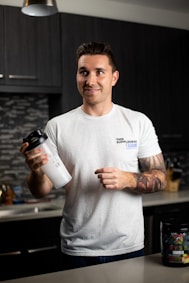 A person with tattoos on their arms is standing in a modern kitchen holding a white shaker cup. They are wearing a casual white t-shirt with a printed logo. The kitchen has dark cabinets and a backsplash featuring a mosaic tile pattern. A container of dietary supplements is placed on the counter.