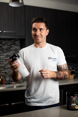 A person with tattoos on their arms is standing in a modern kitchen holding a white shaker cup. They are wearing a casual white t-shirt with a printed logo. The kitchen has dark cabinets and a backsplash featuring a mosaic tile pattern. A container of dietary supplements is placed on the counter.