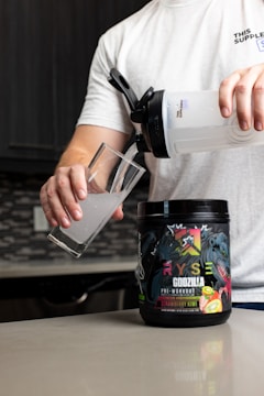 A person is preparing a drink using a shaker bottle and a pre-workout supplement labeled 'Godzilla Strawberry Kiwi'. The person is pouring the liquid into a clear glass on a kitchen counter.