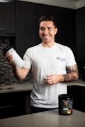 A smiling person stands in a kitchen holding a protein shaker in one hand. They wear a white t-shirt with text on it and have tattoos on their arm. In front of them on the countertop is a container of a dietary supplement. The kitchen has dark cabinets and a tiled backsplash.