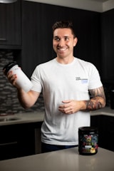 A smiling person stands in a kitchen holding a protein shaker in one hand. They wear a white t-shirt with text on it and have tattoos on their arm. In front of them on the countertop is a container of a dietary supplement. The kitchen has dark cabinets and a tiled backsplash.