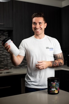 A smiling person stands in a kitchen holding a protein shaker in one hand. They wear a white t-shirt with text on it and have tattoos on their arm. In front of them on the countertop is a container of a dietary supplement. The kitchen has dark cabinets and a tiled backsplash.
