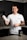 A smiling person stands in a kitchen holding a protein shaker in one hand. They wear a white t-shirt with text on it and have tattoos on their arm. In front of them on the countertop is a container of a dietary supplement. The kitchen has dark cabinets and a tiled backsplash.