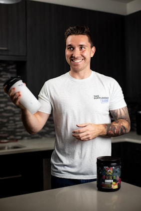A smiling person stands in a kitchen holding a protein shaker in one hand. They wear a white t-shirt with text on it and have tattoos on their arm. In front of them on the countertop is a container of a dietary supplement. The kitchen has dark cabinets and a tiled backsplash.