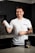 A smiling person stands in a kitchen holding a protein shaker in one hand. They wear a white t-shirt with text on it and have tattoos on their arm. In front of them on the countertop is a container of a dietary supplement. The kitchen has dark cabinets and a tiled backsplash.