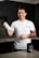 A smiling person stands in a kitchen holding a protein shaker in one hand. They wear a white t-shirt with text on it and have tattoos on their arm. In front of them on the countertop is a container of a dietary supplement. The kitchen has dark cabinets and a tiled backsplash.