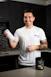 A smiling person stands in a kitchen holding a protein shaker in one hand. They wear a white t-shirt with text on it and have tattoos on their arm. In front of them on the countertop is a container of a dietary supplement. The kitchen has dark cabinets and a tiled backsplash.