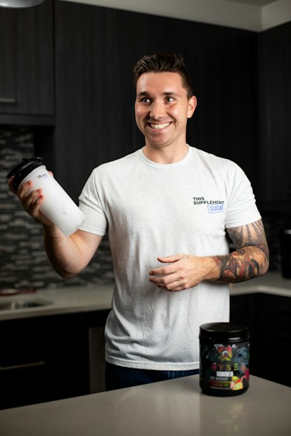 A smiling person stands in a kitchen holding a protein shaker in one hand. They wear a white t-shirt with text on it and have tattoos on their arm. In front of them on the countertop is a container of a dietary supplement. The kitchen has dark cabinets and a tiled backsplash.