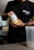 A person is holding a container of sports supplement powder in one hand while the other hand is on the lid. The person is wearing a black shirt with a humorous message and has tattoos on the arms. A blender bottle sits on the table in front.