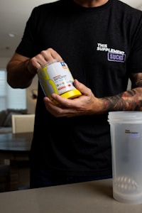 A person is holding a container of sports supplement powder in one hand while the other hand is on the lid. The person is wearing a black shirt with a humorous message and has tattoos on the arms. A blender bottle sits on the table in front.