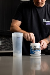 A person is preparing a pre-workout supplement in a kitchen setting. They are wearing a black t-shirt with text on it and scooping powder from a container labeled 'PRE-WORKOUT'. A clear shaker bottle is placed on the counter next to them. The background includes a modern kitchen with dark cabinetry and a tiled backsplash.