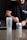 A person is preparing a pre-workout supplement in a kitchen setting. They are wearing a black t-shirt with text on it and scooping powder from a container labeled 'PRE-WORKOUT'. A clear shaker bottle is placed on the counter next to them. The background includes a modern kitchen with dark cabinetry and a tiled backsplash.