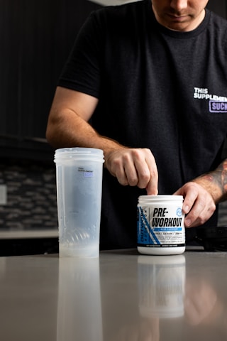 A person is preparing a pre-workout supplement in a kitchen setting. They are wearing a black t-shirt with text on it and scooping powder from a container labeled 'PRE-WORKOUT'. A clear shaker bottle is placed on the counter next to them. The background includes a modern kitchen with dark cabinetry and a tiled backsplash.