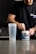 A person is preparing a pre-workout supplement in a kitchen setting. They are wearing a black t-shirt with text on it and scooping powder from a container labeled 'PRE-WORKOUT'. A clear shaker bottle is placed on the counter next to them. The background includes a modern kitchen with dark cabinetry and a tiled backsplash.