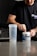 A person is preparing a pre-workout supplement in a kitchen setting. They are wearing a black t-shirt with text on it and scooping powder from a container labeled 'PRE-WORKOUT'. A clear shaker bottle is placed on the counter next to them. The background includes a modern kitchen with dark cabinetry and a tiled backsplash.