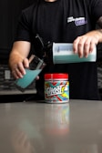 A person holds a shaker bottle and pours a blue liquid into a glass. The shaker is positioned over a colorful tub labeled 'Ghost Legend' which sits on a countertop. The person is wearing a black t-shirt with text on the chest.