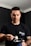 A person wearing a black t-shirt with a humorous design 'THIS SUPPLEMENT SUCKS' stands indoors while holding a scoop of powder above an open container. The individual appears focused and is in a room with dark walls.