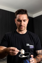 A person wearing a black t-shirt with a humorous design 'THIS SUPPLEMENT SUCKS' stands indoors while holding a scoop of powder above an open container. The individual appears focused and is in a room with dark walls.