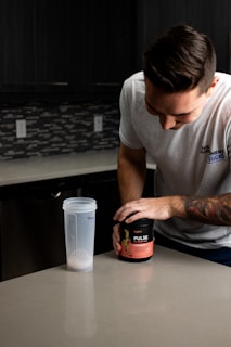 A person in a gray T-shirt is preparing a supplement drink in a modern kitchen. The individual is holding a container labeled 'Legion Pulse' next to a clear shaker bottle with some powder inside. The countertop is clean and smooth, and the background features dark cabinets and a tiled backsplash.