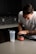 A person in a gray T-shirt is preparing a supplement drink in a modern kitchen. The individual is holding a container labeled 'Legion Pulse' next to a clear shaker bottle with some powder inside. The countertop is clean and smooth, and the background features dark cabinets and a tiled backsplash.