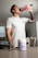 A person with tattoos is drinking from a pink shaker bottle in a kitchen. There is a tub labeled 'Thavage Pre-Workout' on a counter along with an empty glass. The setting appears to be modern with neutral tones and soft lighting.