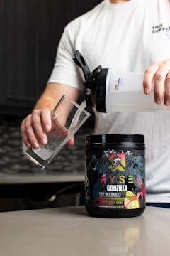 A person in a white shirt is pouring a drink from a large shaker bottle into a glass. A container of RYSE Godzilla pre-workout supplement with strawberry kiwi flavor is placed on the counter.