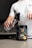 A person in a white shirt is pouring a drink from a large shaker bottle into a glass. A container of RYSE Godzilla pre-workout supplement with strawberry kiwi flavor is placed on the counter.