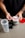 A person with tattooed arms is holding a scoop of white powder above an open container of Ghost branded supplement. The container is placed on a light-colored table along with a partially assembled shaker bottle and a red lid.