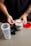 A person with tattooed arms is holding a scoop of white powder above an open container of Ghost branded supplement. The container is placed on a light-colored table along with a partially assembled shaker bottle and a red lid.