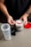 A person with tattooed arms is holding a scoop of white powder above an open container of Ghost branded supplement. The container is placed on a light-colored table along with a partially assembled shaker bottle and a red lid.
