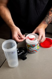 A person with tattooed arms is holding a scoop of white powder above an open container of Ghost branded supplement. The container is placed on a light-colored table along with a partially assembled shaker bottle and a red lid.