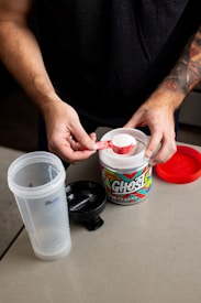 A person with tattooed arms is holding a scoop of white powder above an open container of Ghost branded supplement. The container is placed on a light-colored table along with a partially assembled shaker bottle and a red lid.