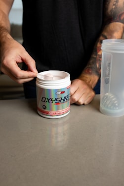 A person is scooping powder from a container labeled 'Oxyshred' with the flavor 'Juicy Watermelon' indicated. The person is wearing a dark shirt and has tattoos on their arm. A translucent shaker bottle is visible on the countertop next to the container.