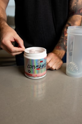 A person is scooping powder from a container labeled 'Oxyshred' with the flavor 'Juicy Watermelon' indicated. The person is wearing a dark shirt and has tattoos on their arm. A translucent shaker bottle is visible on the countertop next to the container.