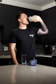 A person stands in a kitchen drinking from a shaker bottle filled with a light-colored liquid. They are wearing a black t-shirt and have a tattooed arm. On the counter, there is an empty glass and a container labeled 'Wrecked', which appears to be a supplement or pre-workout product. The kitchen has dark cabinets and a tiled backsplash.