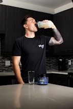 A person stands in a kitchen drinking from a shaker bottle filled with a light-colored liquid. They are wearing a black t-shirt and have a tattooed arm. On the counter, there is an empty glass and a container labeled 'Wrecked', which appears to be a supplement or pre-workout product. The kitchen has dark cabinets and a tiled backsplash.