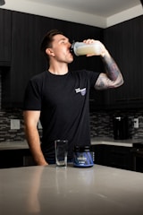 A person stands in a kitchen drinking from a shaker bottle filled with a light-colored liquid. They are wearing a black t-shirt and have a tattooed arm. On the counter, there is an empty glass and a container labeled 'Wrecked', which appears to be a supplement or pre-workout product. The kitchen has dark cabinets and a tiled backsplash.
