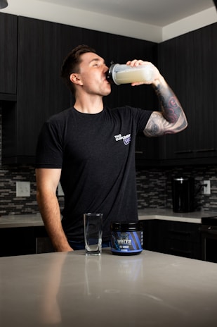 A person stands in a kitchen drinking from a shaker bottle filled with a light-colored liquid. They are wearing a black t-shirt and have a tattooed arm. On the counter, there is an empty glass and a container labeled 'Wrecked', which appears to be a supplement or pre-workout product. The kitchen has dark cabinets and a tiled backsplash.