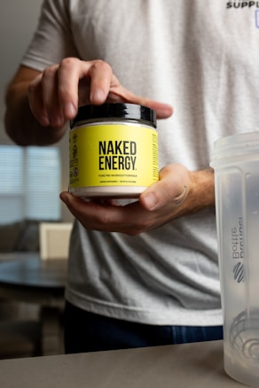 A person holding a container labeled 'Naked Energy' against a blurred interior background. The container has a bright yellow label with black text.