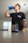 A person is drinking from a blue shaker bottle in a room with a container of pre-workout supplement prominently displayed in the foreground on a table. The person is wearing a dark t-shirt and has a tattooed arm.