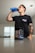 A person is drinking from a blue shaker bottle in a room with a container of pre-workout supplement prominently displayed in the foreground on a table. The person is wearing a dark t-shirt and has a tattooed arm.