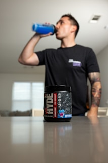 A person is drinking from a blue shaker bottle in a room with a container of pre-workout supplement prominently displayed in the foreground on a table. The person is wearing a dark t-shirt and has a tattooed arm.
