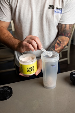 A person is scooping a powdered supplement from a container labeled 'Naked Energy' into a shaker cup. The individual is wearing a light gray t-shirt with a print on it. There are tattoos visible on the person's arm. The container is bright yellow with black lettering, and the shaker cup is translucent with some text on it.