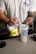 A person is scooping a powdered supplement from a container labeled 'Naked Energy' into a shaker cup. The individual is wearing a light gray t-shirt with a print on it. There are tattoos visible on the person's arm. The container is bright yellow with black lettering, and the shaker cup is translucent with some text on it.