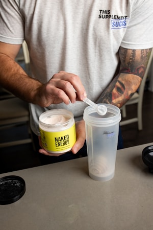 A person is scooping a powdered supplement from a container labeled 'Naked Energy' into a shaker cup. The individual is wearing a light gray t-shirt with a print on it. There are tattoos visible on the person's arm. The container is bright yellow with black lettering, and the shaker cup is translucent with some text on it.