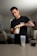 A person is preparing a drink using a supplement powder. The individual is standing at a kitchen counter pouring the contents from a yellow container into a large clear shaker bottle. The setting appears to be a modern kitchen or living space, with a round table and chairs in the background. The person's arm tattoos and casual black t-shirt are also visible.