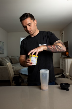 A person is preparing a drink using a supplement powder. The individual is standing at a kitchen counter pouring the contents from a yellow container into a large clear shaker bottle. The setting appears to be a modern kitchen or living space, with a round table and chairs in the background. The person's arm tattoos and casual black t-shirt are also visible.