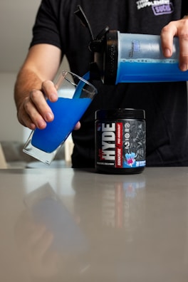 A person is pouring a vibrant blue liquid into a tall glass from a shaker bottle. Next to the glass on the table, there is a container labeled 'Hyde pre-workout' with graphics and text. The person is wearing a dark t-shirt, and the setting appears to be a kitchen or dining area with a reflective surface.