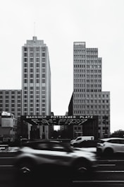 Two modern, rectangular buildings tower over a busy urban street. In front of them, the Bahnhof Potsdamer Platz entrance is visible, with blurred cars in motion adding a dynamic contrast to the otherwise still architecture. The image is monochromatic, giving it a classic, timeless feel.