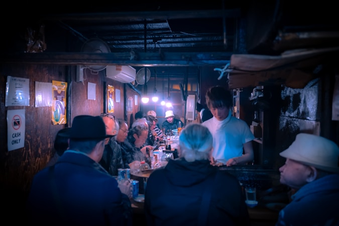 A small, dimly lit restaurant or bar with several people seated along a counter, engaging in conversation and dining. The atmosphere is cozy and intimate, with low lighting emanating from lamps above the counter. The walls are adorned with various signs and posters, including a 'CASH ONLY' notice. The server is interacting with patrons, creating a sense of community.